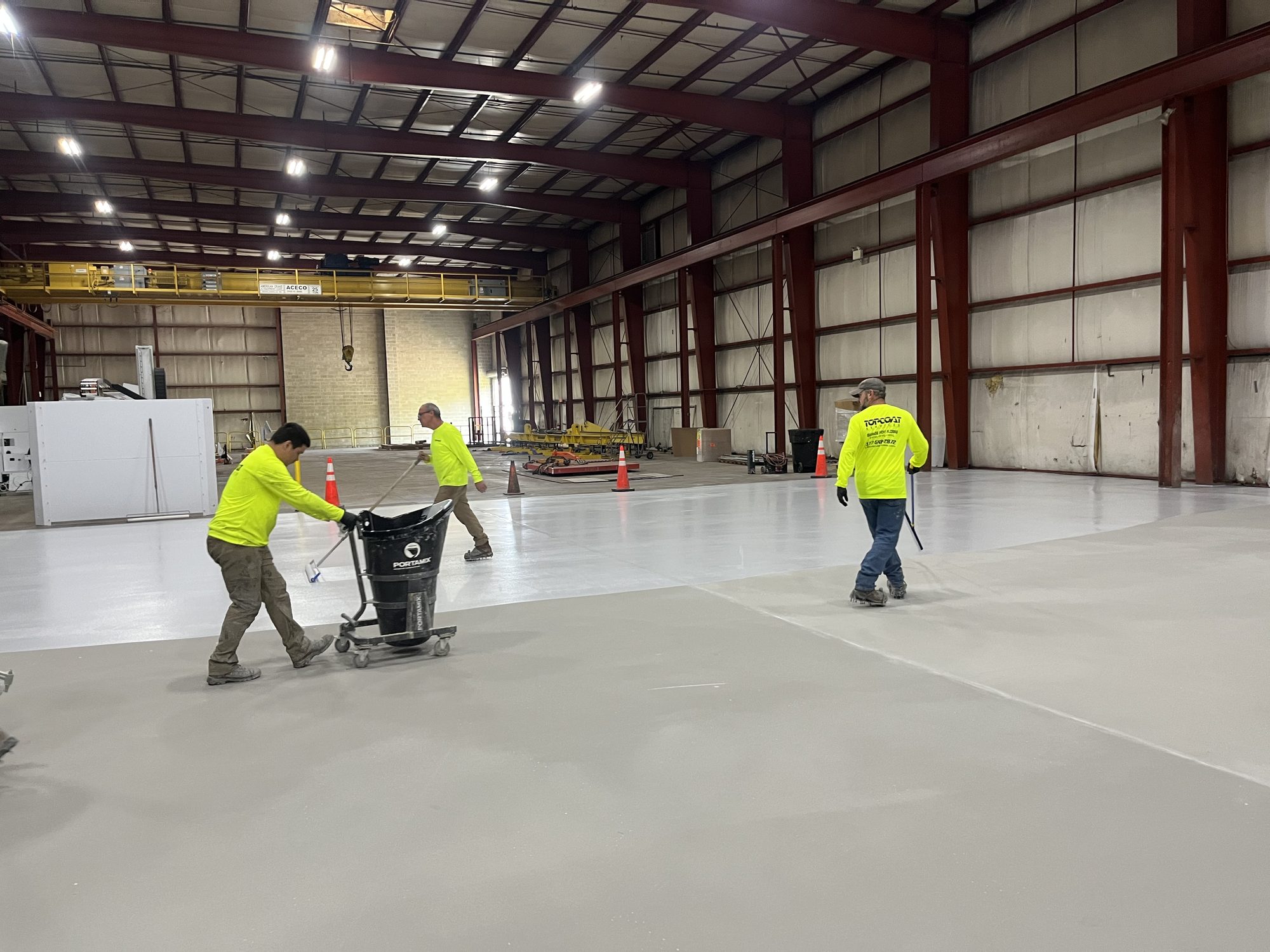Industrial Flooring: Best Options for High-Traffic Facilities 8 Topcoat crew applying a coat across the fabrication bay with part of the floor already coated, American Steel and Aluminum, Auburn, MA